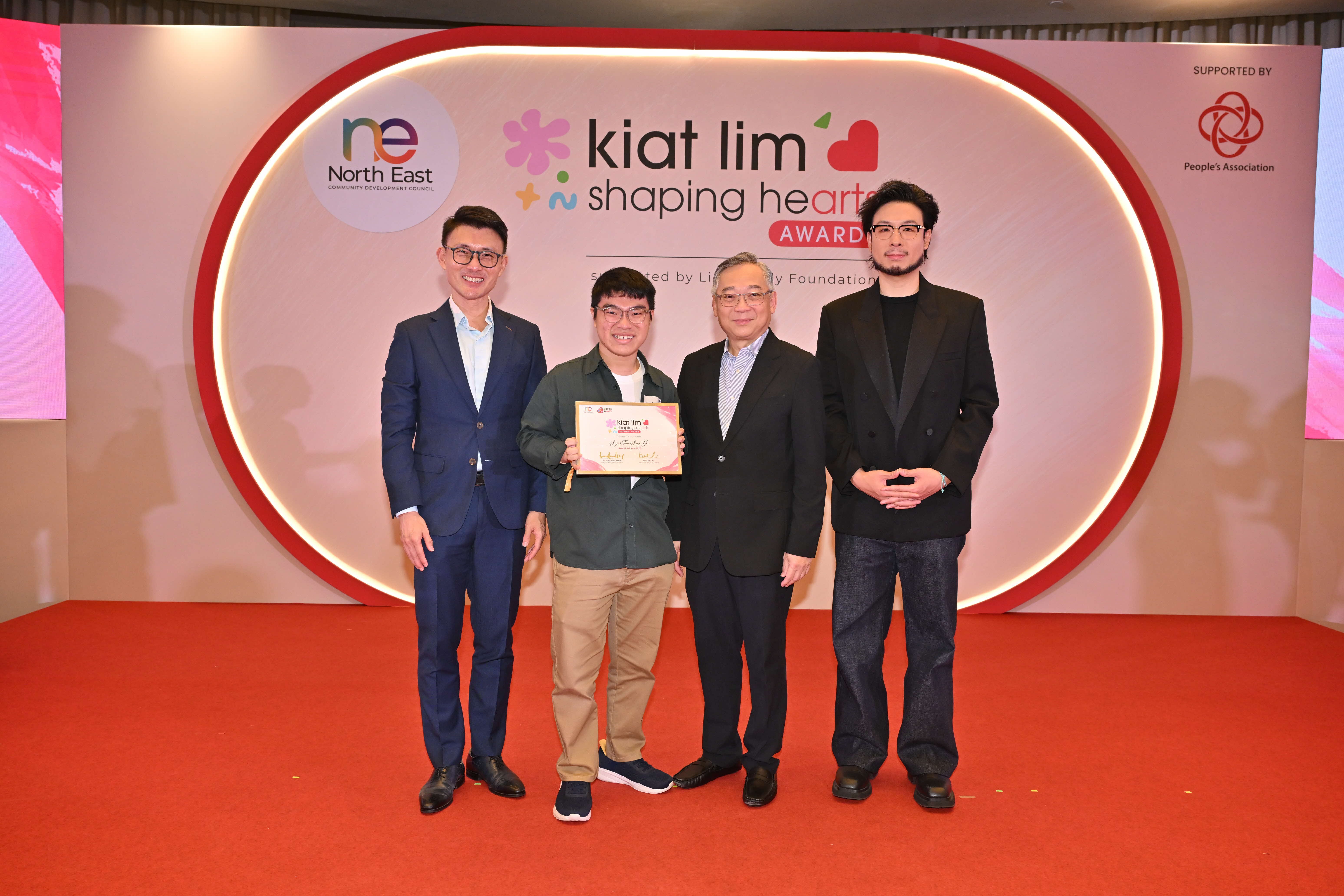 Award recipient standing on stage with the presenter, holding their certificate and posing for a group photo during the award ceremony, with a backdrop reading ‘Kiat Lim-Shaping Hearts Awards'.